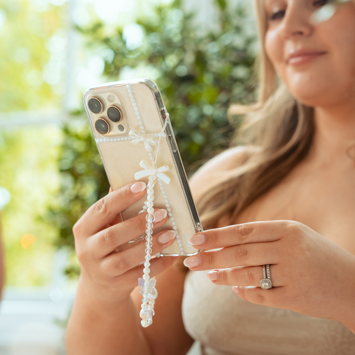 Woman with manicured nails holds a clear phone case with pearl bow details, her engagement ring visible.