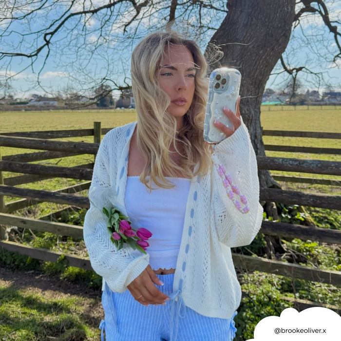 Woman holding phone with pink daisy crochet flower phone strap, standing outdoors near a fence and field.