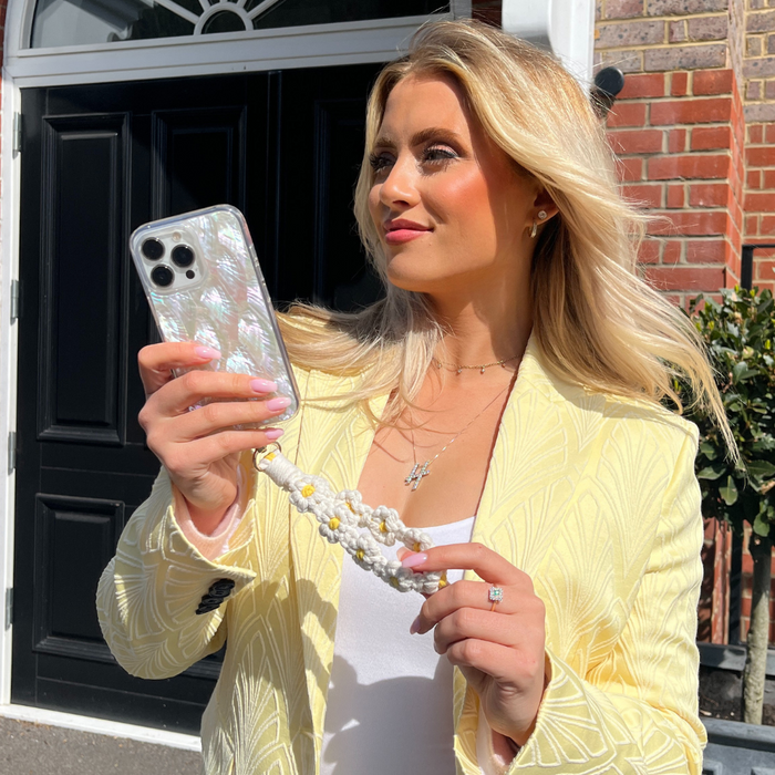 Woman holding phone with white daisy crochet flower phone strap by a black door.
