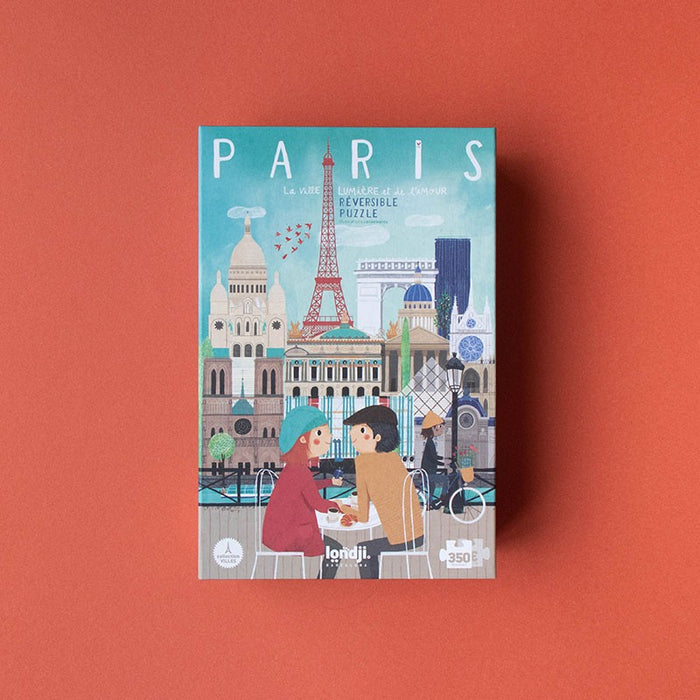 A puzzle box with an illustration of a couple sitting in front of a puzzle board with Parisian landmarks like the Eiffel Tower and the Louvre.