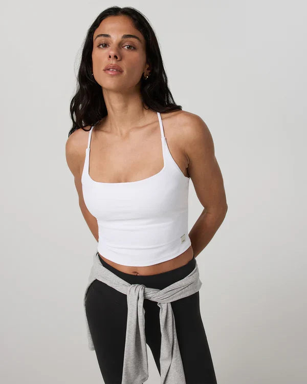 Woman wearing a white Vuori - AllTheFeels Strappy Back top and black leggings with a gray towel over her shoulder on a light gray background