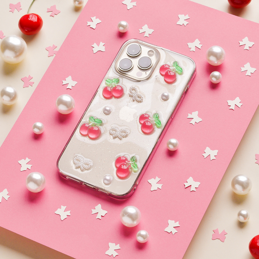 Coconut Lane Cherries & Bows iPhone 15 case on pink paper, surrounded by pearls and star-shaped confetti.