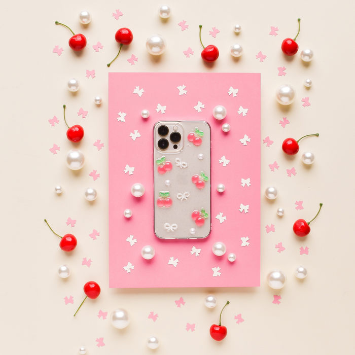Coconut Lane phone case with cherry and bow pattern on pink paper, surrounded by cherries, pearls, and confetti.