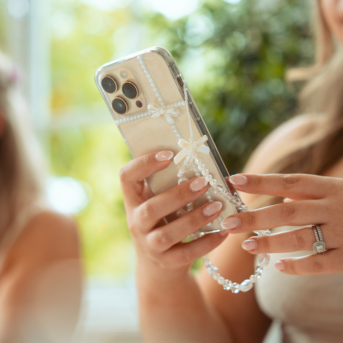 Clear iPhone 15 case with pearl and bow decorations, held by a woman with manicured nails.
