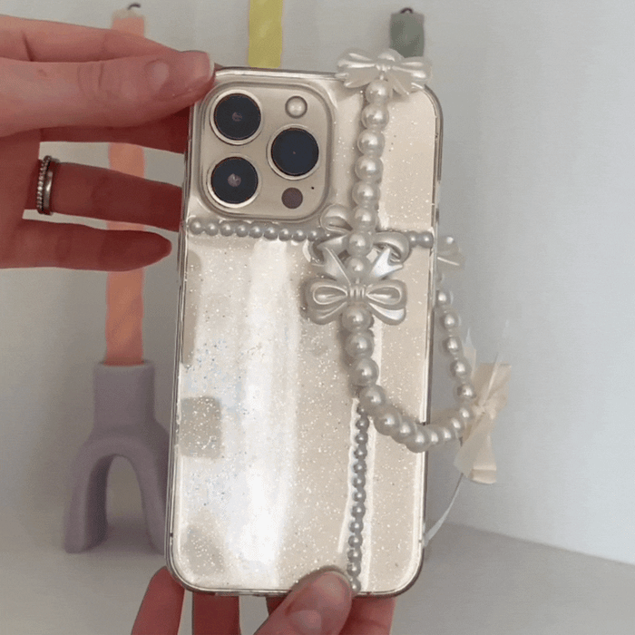 Hand holding a clear iPhone case with pearl bow design; colorful candles softly glowing in the background.