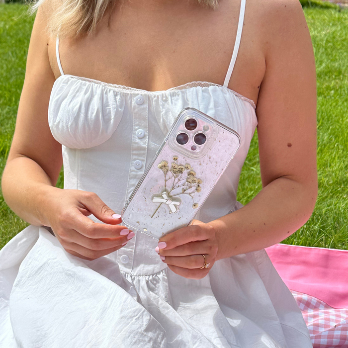 Woman in a white dress on grass holds a clear iPhone 15 case with dried flowers; pink blanket nearby.