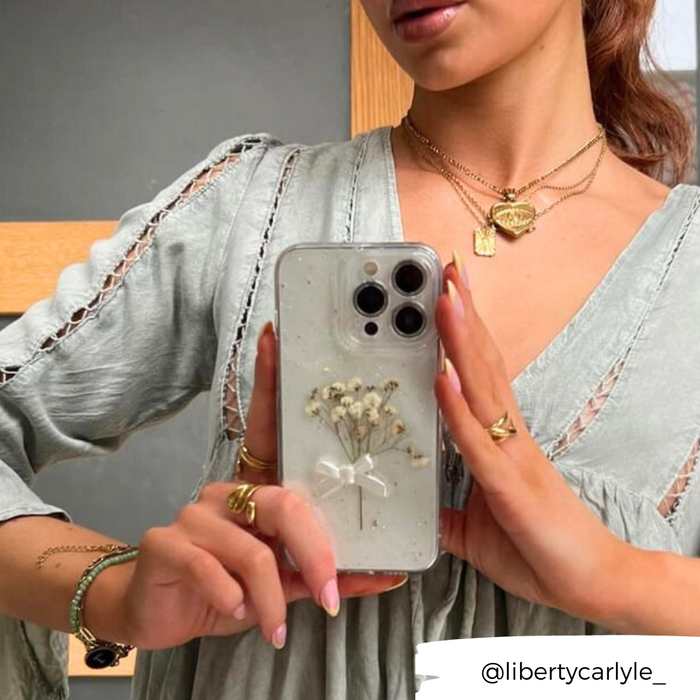 Woman in pale dress holding a clear phone case with dried flower design, white bow, and gold jewelry visible.
