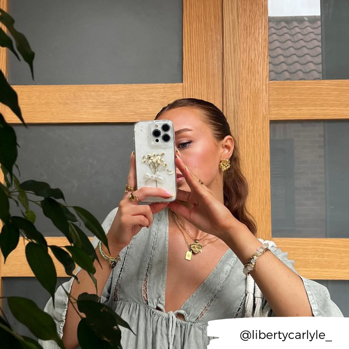 Woman holding a clear phone case with dried flower design, wearing layered necklaces and rings, taking a mirror selfie.