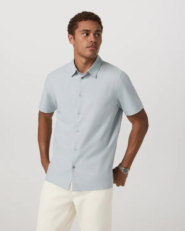 Man wearing a light blue Vuori - Short Sleeve Manhattan Button Down shirt and white pants on a plain background