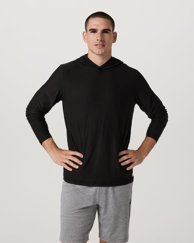 Man wearing a Vuori black Strato hoodie and gray shorts on a light gray background