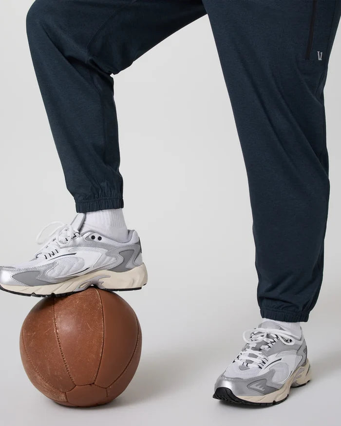 Person wearing Vuori navy pants and gray sneakers stepping on a brown medicine ball against a light background