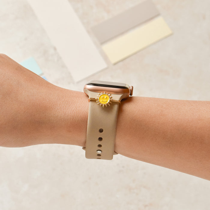 Gold Smiley Sun charm on beige silicone Coconut Lane watch strap, with additional watch band accessories in the background.