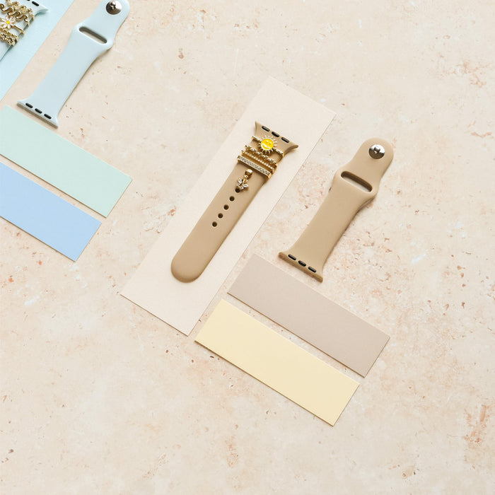 Beige silicone watch straps with a gold smiley sun charm, displayed on a light marble surface.